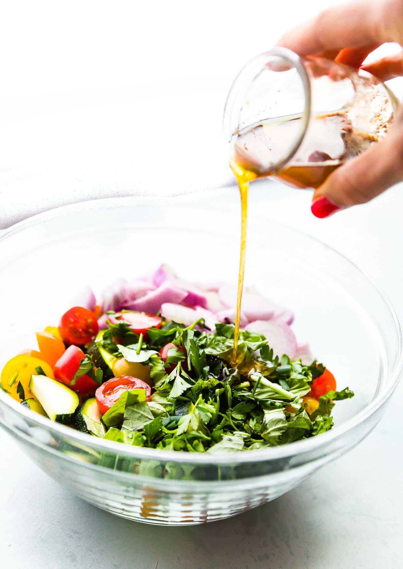 Woman's hand pouring salad dressing into a bowl of chopped vegetables and herbs