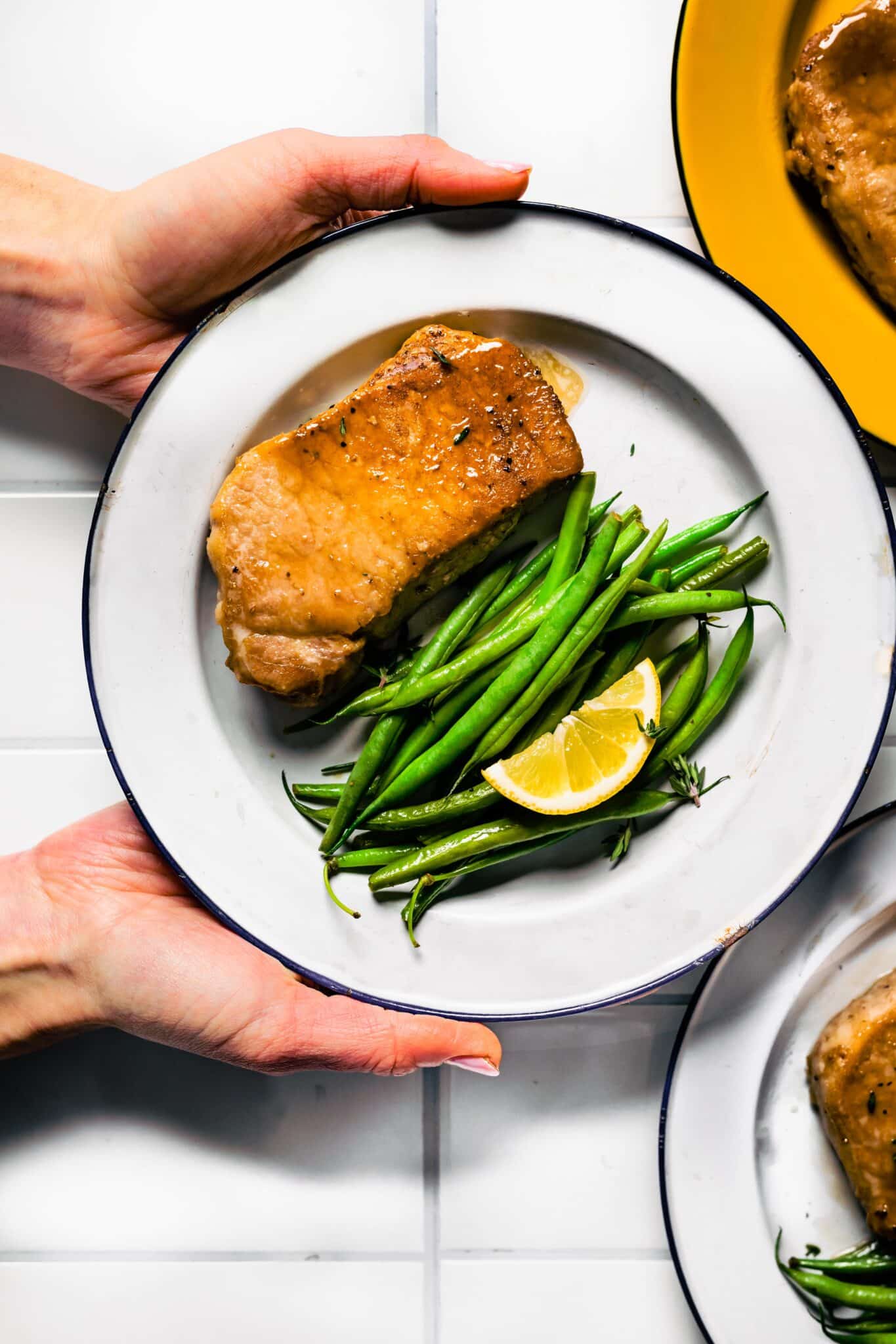 A woman holding a white plate with a glazed pork chop and greens beans.