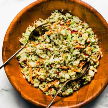 Two metal serving spoons in apple slaw in a wooden bowl.