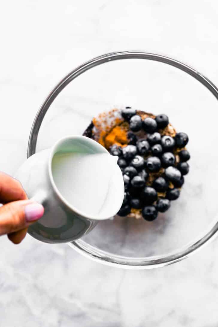 woman pouring milk into a prep bowl of blueberry overnight oats