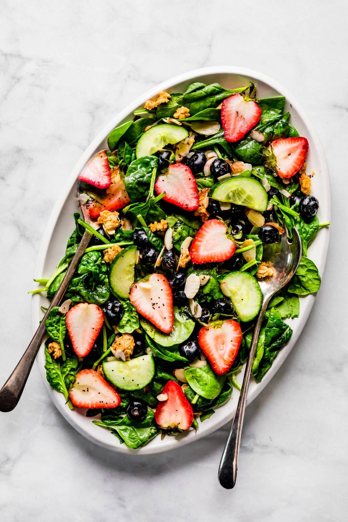 Overhead photo of a strawberry spinach salad on a white platter with salad servers.