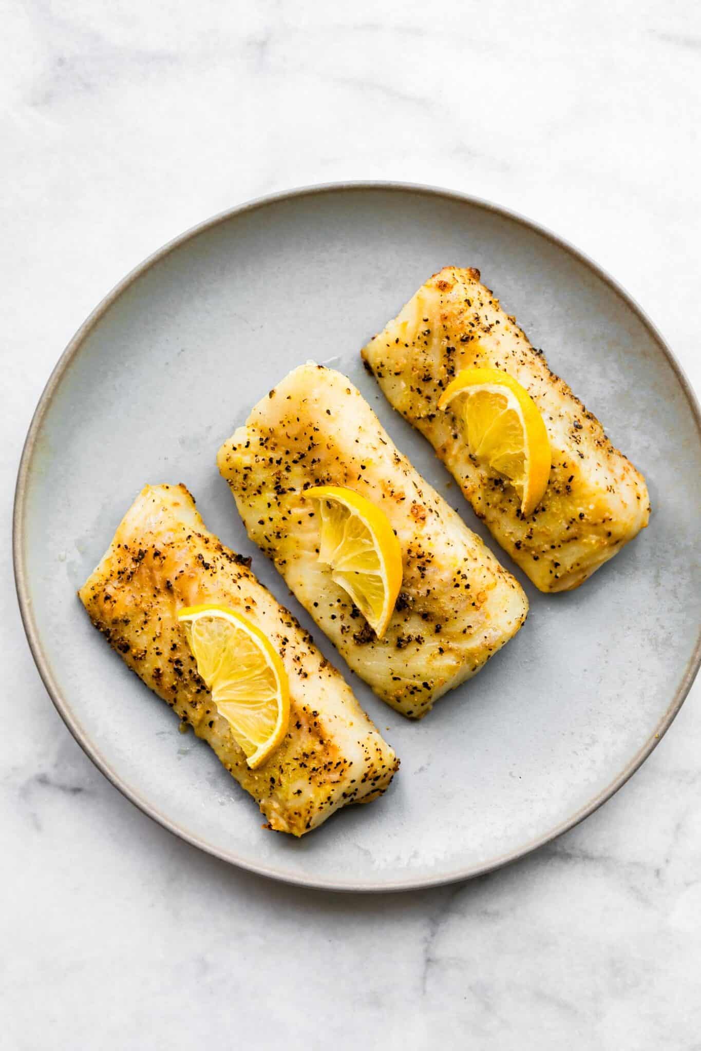 Three cooked fish fillets topped with lemon on a grey plate