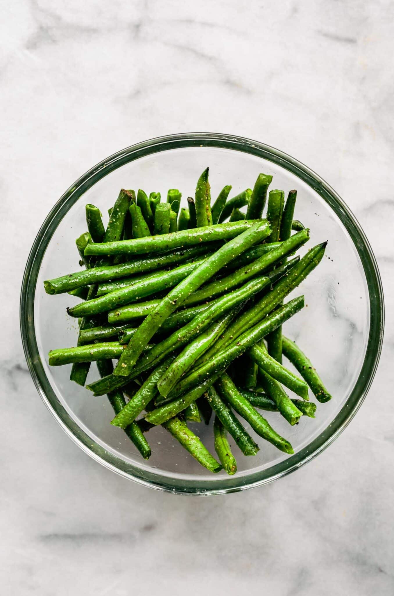 Fresh green beans mixed with seasonings and oil in a glass bowl.