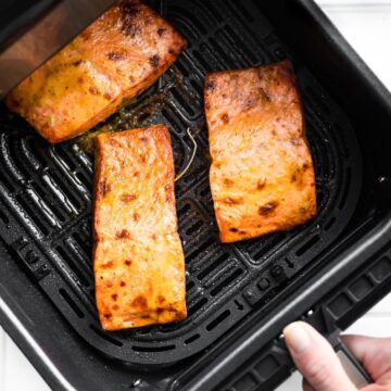 a woman's hand holding the handle of an air fryer basket that is filled with 3 pieces of cooked air fryer salmon topped with creamy mustard sauce