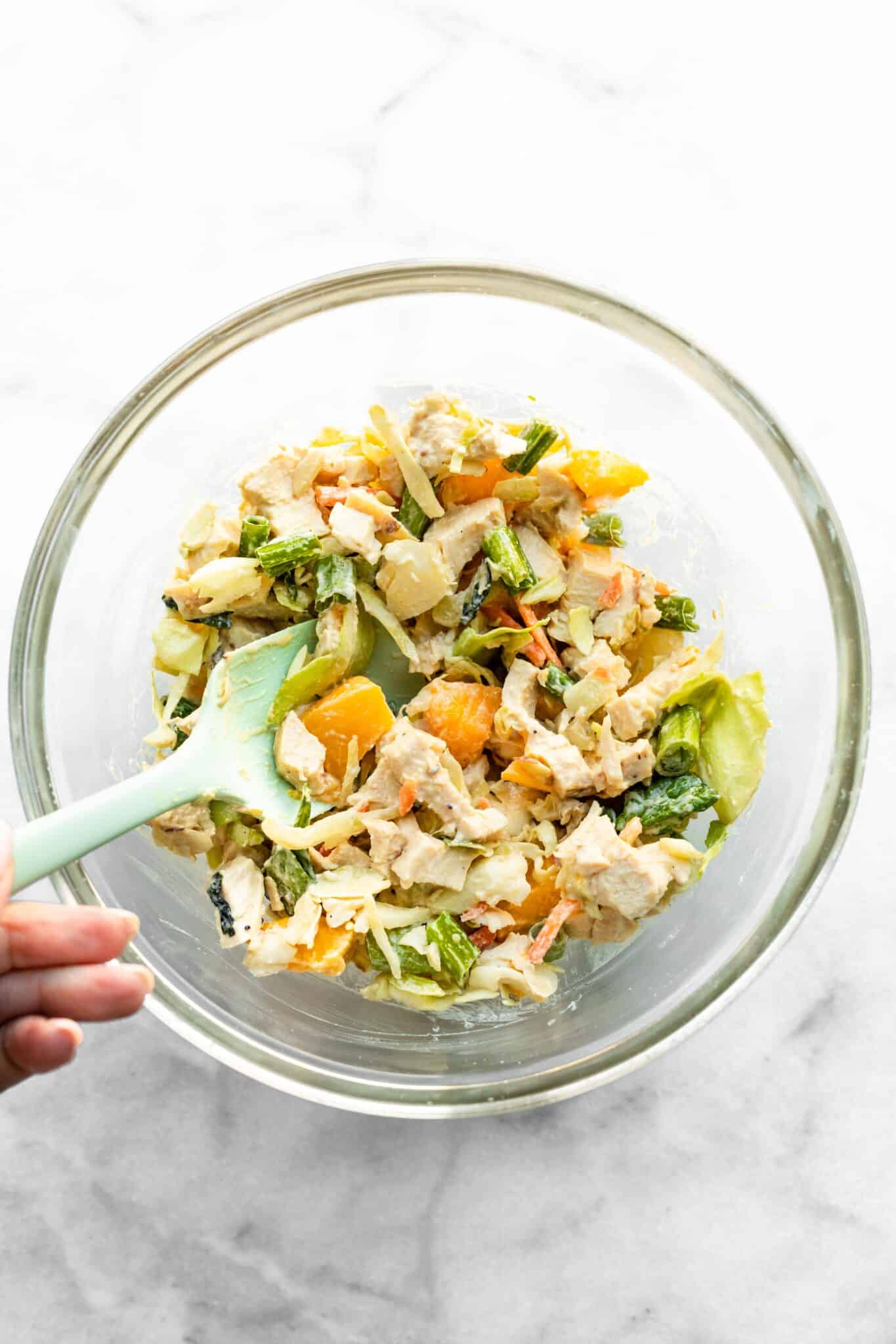 A blue spatula combining the ingredients for Asian chicken salad in a glass bowl,