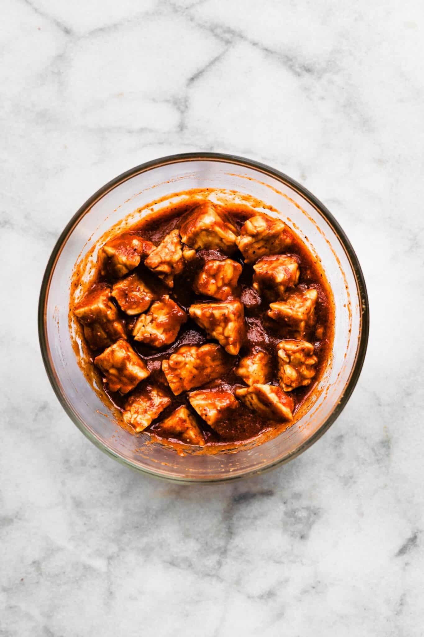 Cubed tempeh marinating in BBQ sauce in a glass bowl.