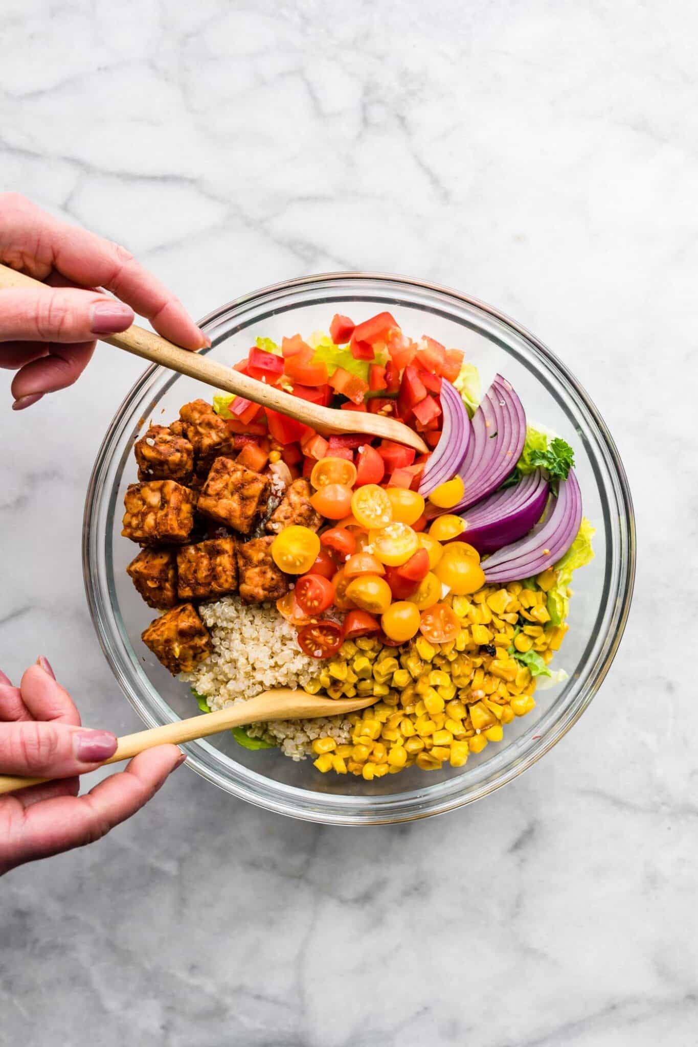 A woman's hands using wooden spoons to toss a BBQ tempeh salad with fresh vegetables.