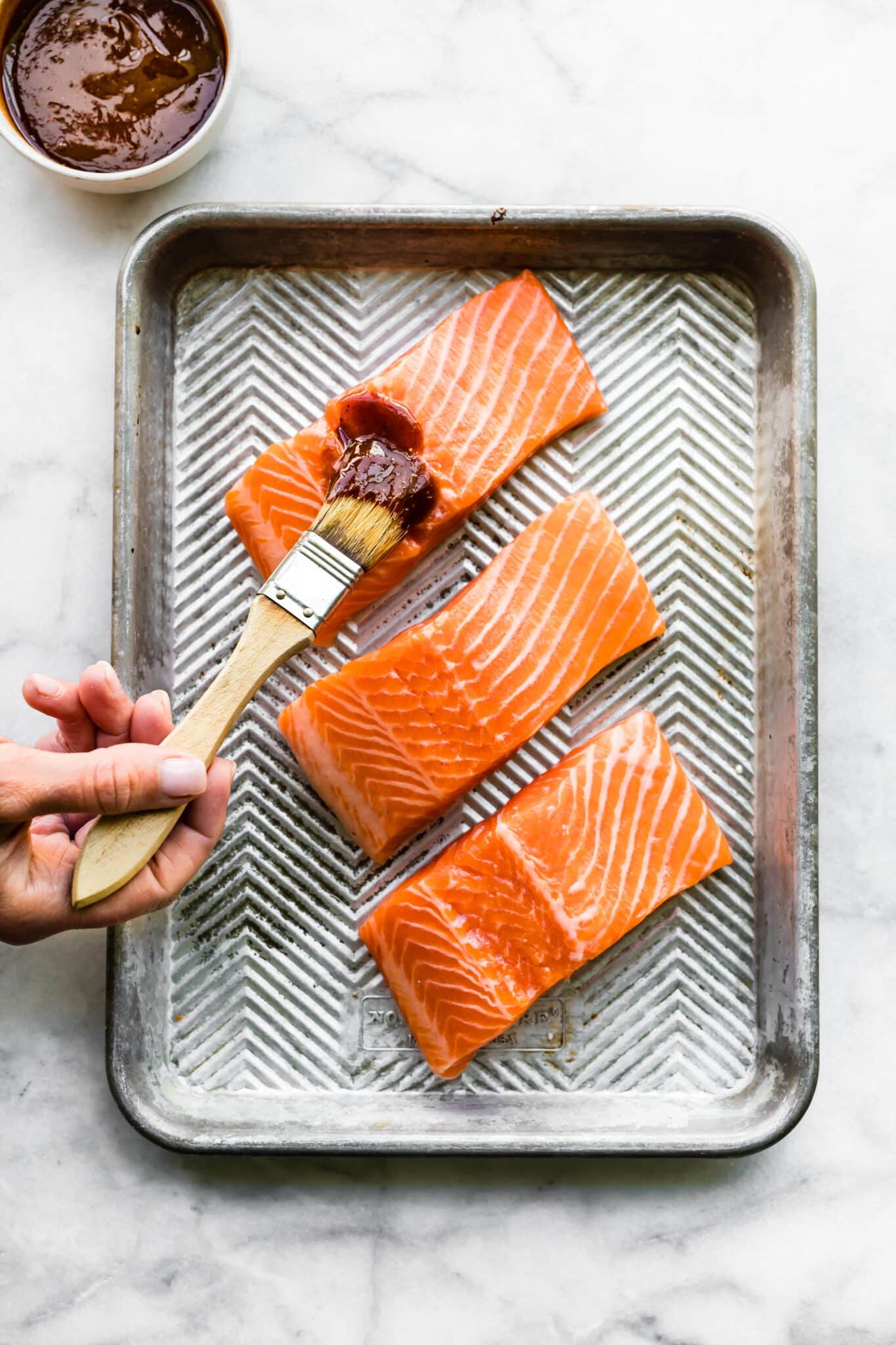 A woman using a brush to coat fresh salmon filets with nightshade free BBQ sauce.