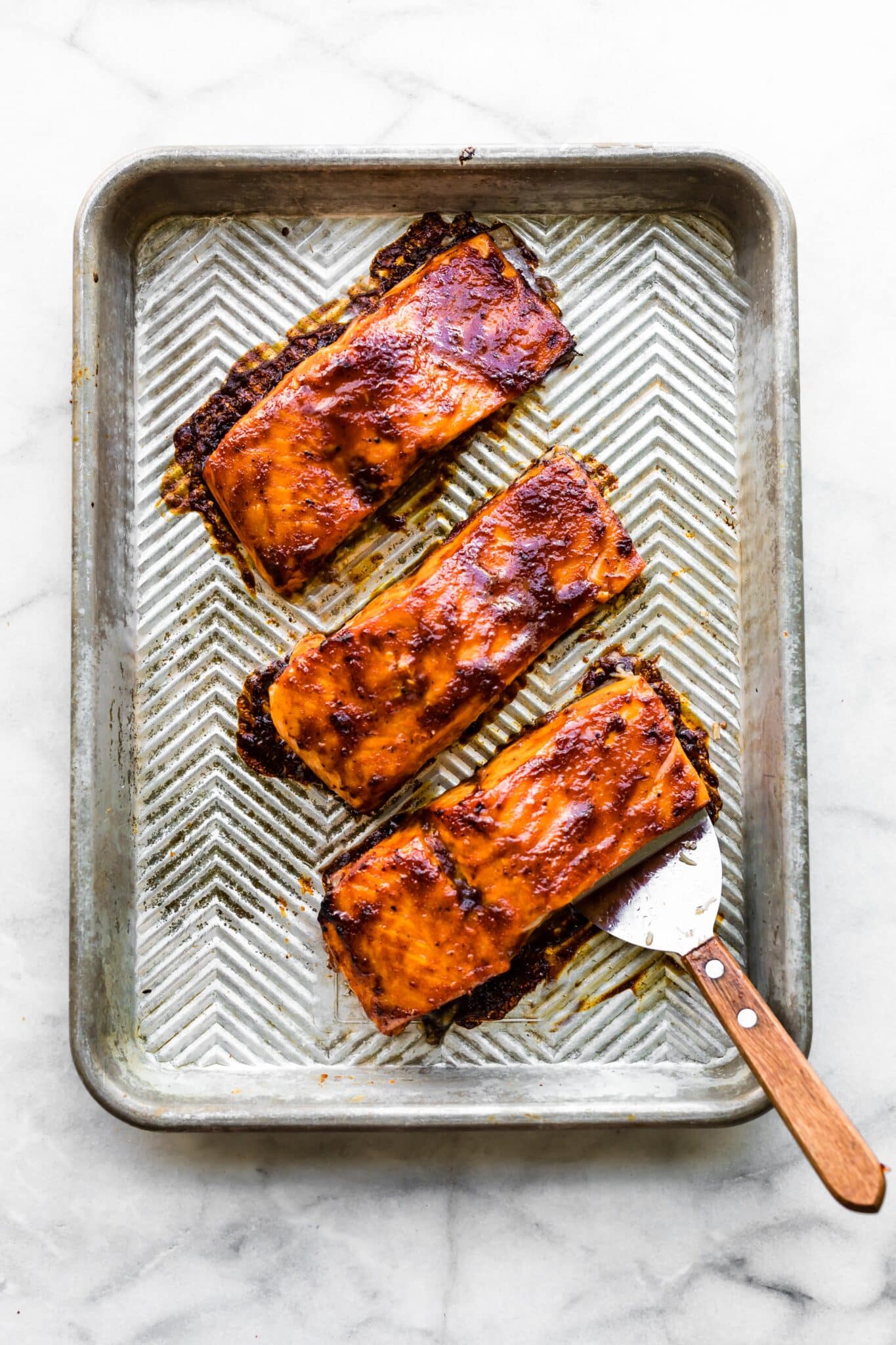 Overhead photo of three oven baked salmon filets on a sheet pan with spatula.