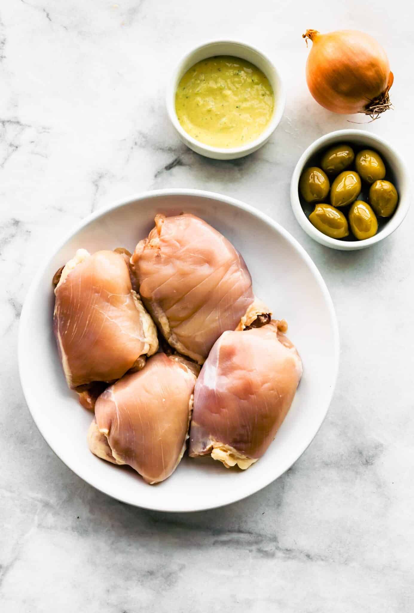 4 raw Chicken Thighs on a plate next an onion, a bowl of Olives and lemon sage