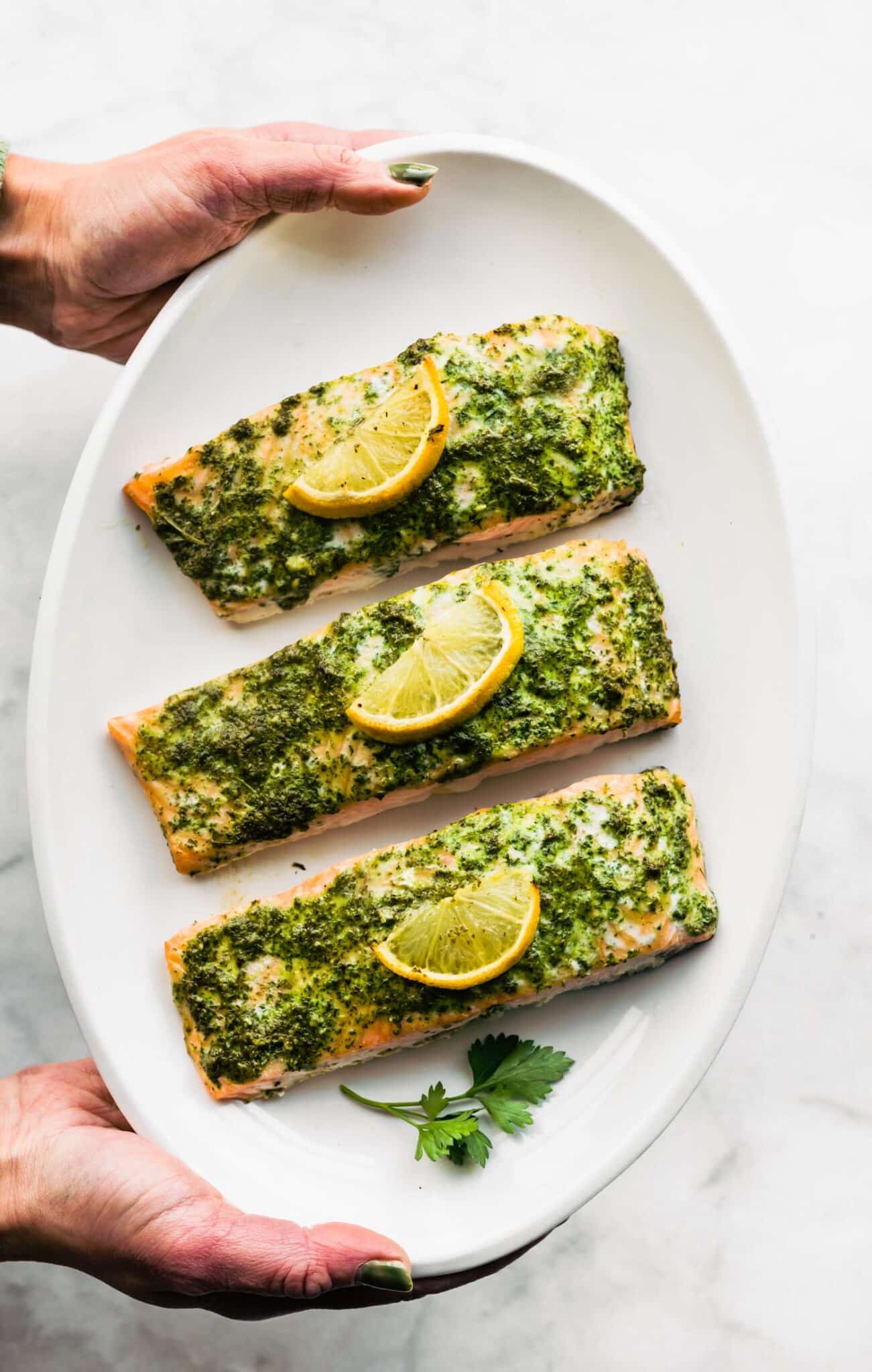 A woman's hands holding a white platter with three salmon filets topped with herb sauce.