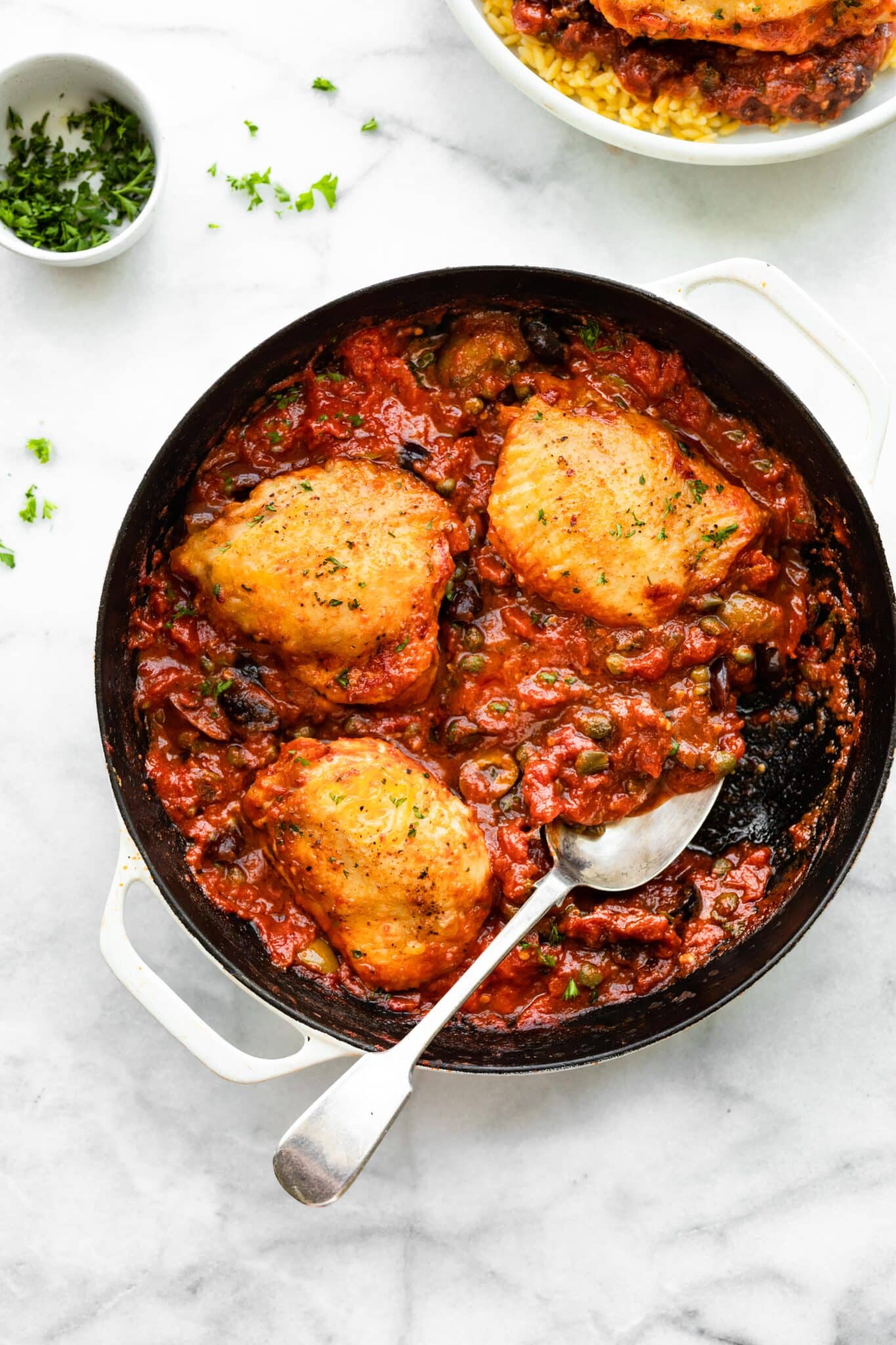 Photo of a spoon in a pan of chicken puttanesca with one chicken thigh removed.
