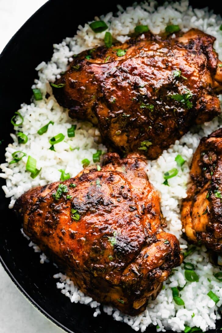close up of 2 Jerk chicken thighs served on a bed of white rice in a black skillet, topped with chopped green onions