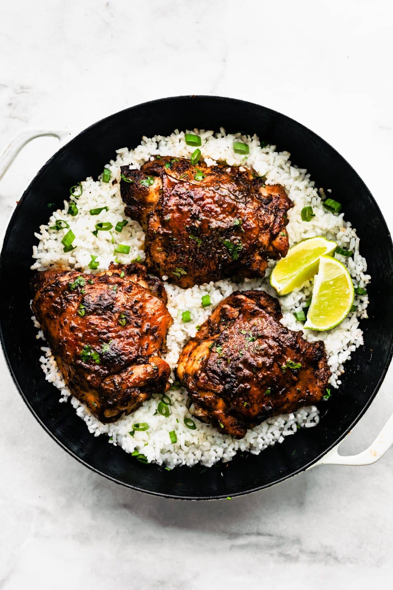 Jerk chicken thighs served on a bed of fluffy white rice in a black skillet, topped with chopped green onions and lime wedges on the side.