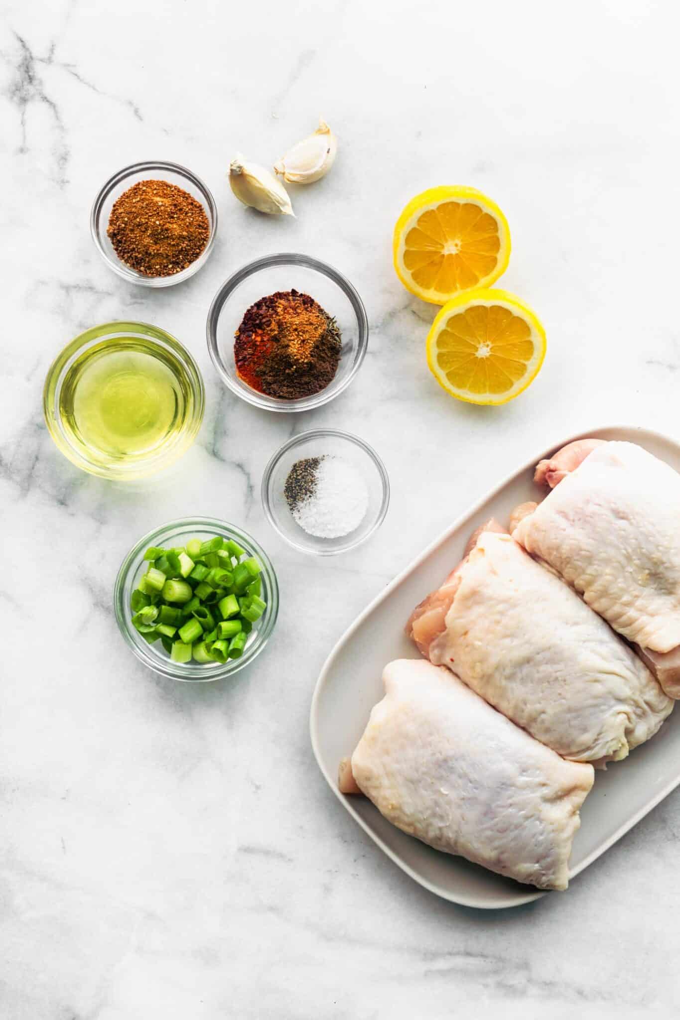 Overhead shot of raw bone-in chicken thighs on a white plate, surrounded by small bowls of jerk seasoning spices, oil, chopped green onions, garlic cloves, salt, pepper, and halved lemons on a marble background.
