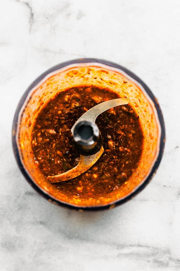 Close-up overhead shot of a blender bowl filled with blended jerk marinade, deep red and flecked with herbs and spices.
