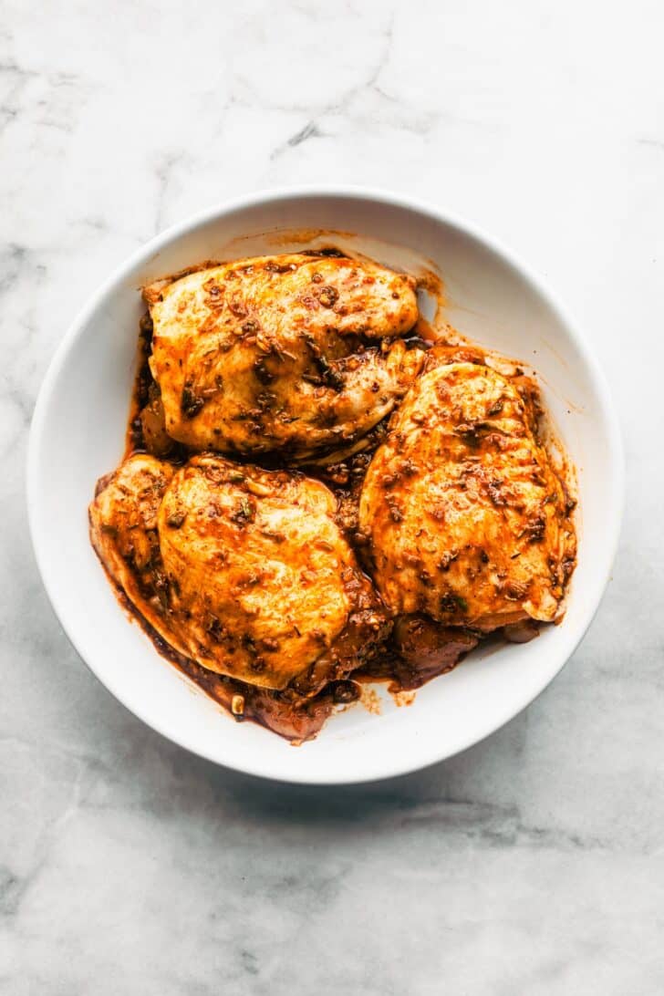 Raw chicken thighs coated in rich jerk marinade, sitting in a white mixing bowl.