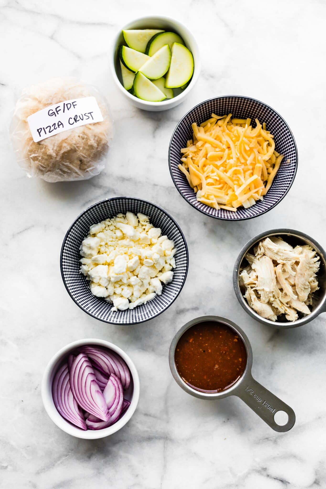 Ingredients in bowls for BBQ chicken pizza on a white marble countertop.