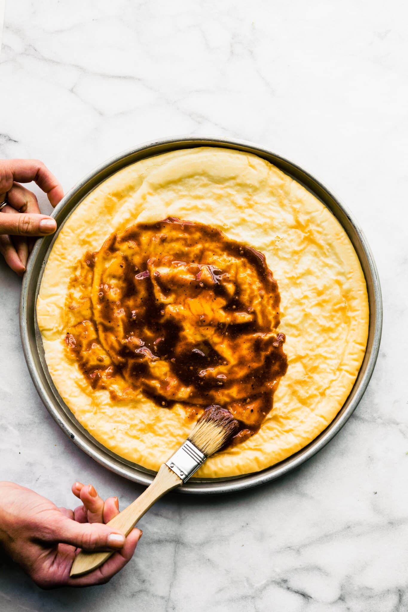 A woman brushing nightshade free BBQ sauce on a parbaked pizza crust.
