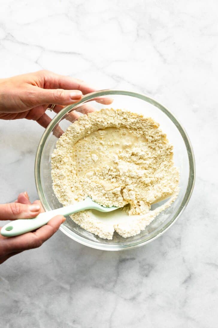 A woman's hands using a silicone spatula to mix wet ingredients into gluten free flour.
