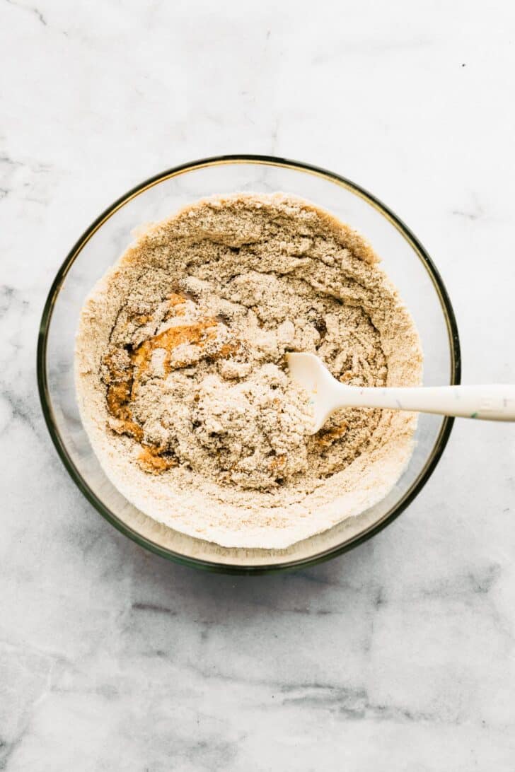 Gluten-free dry ingredients being folded into wet mixture in a clear bowl with a spatula.