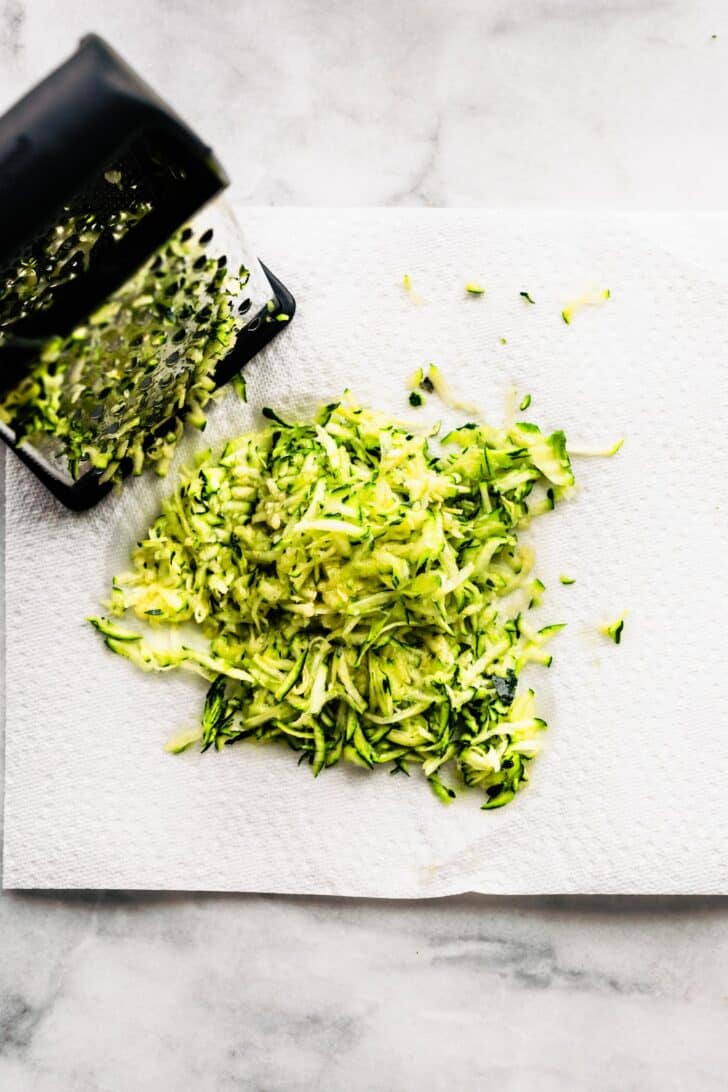 a grater with zucchini in it and grated zucchini on a white paper towel