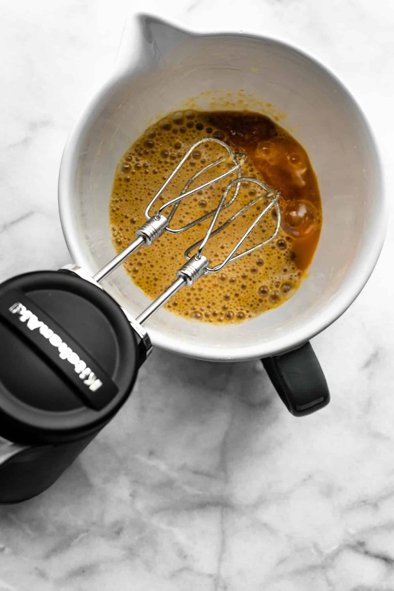 A handheld mixer mixing wet ingredients in bowl.