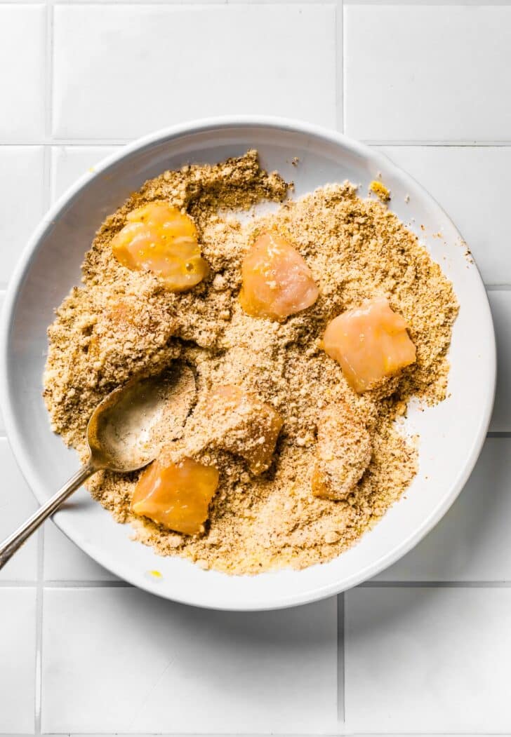et. Chicken pieces being coated in gluten-free breading mixture in a white bowl with a spoon.