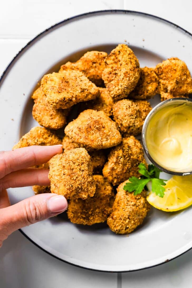 Close-up of a hand holding a crispy gluten-free chicken nugget with a bite taken out, showing juicy white chicken inside.