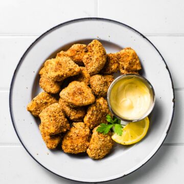 Finished gluten-free chicken nuggets on a white plate with a small dipping bowl of sauce, lemon wedge, and parsley garnish.