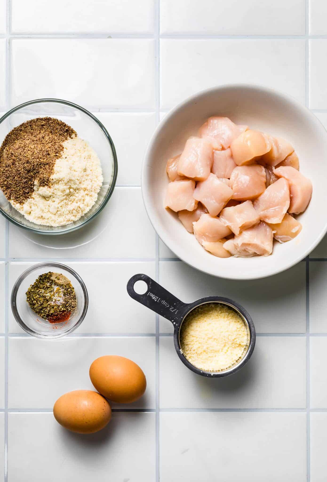 Overhead view of raw chicken pieces in a bowl surrounded by ingredients: almond flour, flax meal, parmesan, spices, and eggs.