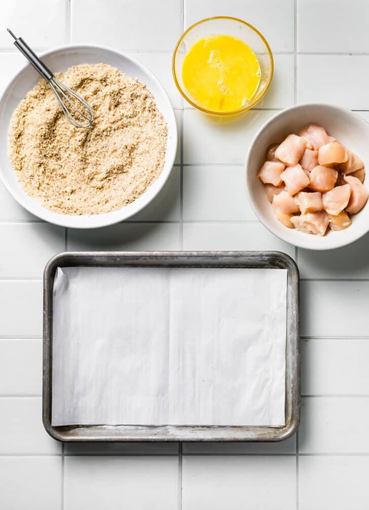 Prep station with whisked eggs, seasoned breading mixture, raw chicken cubes, and a parchment-lined baking sheet.