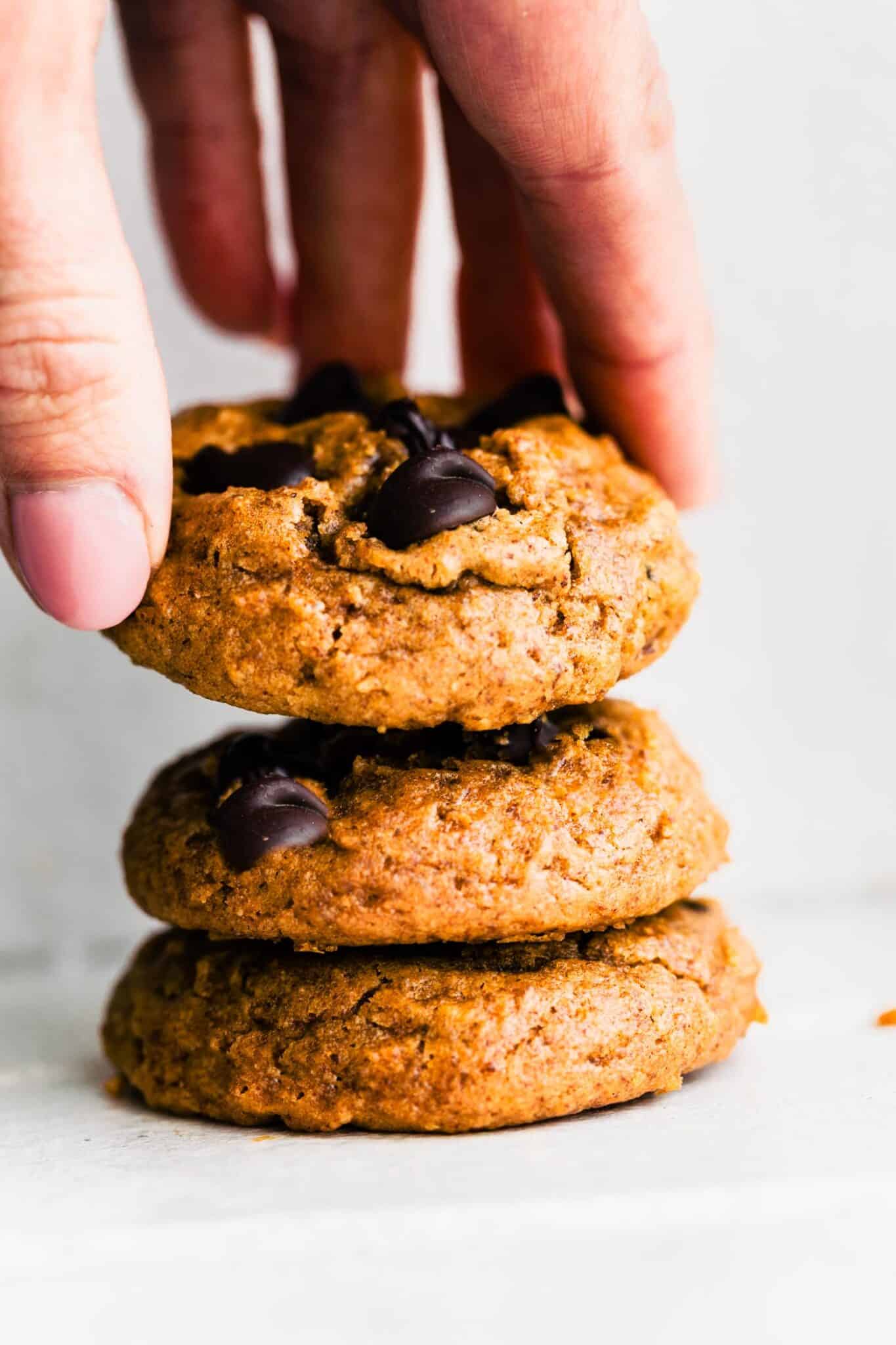Close-up of three pumpkin chocolate chip cookies stacked, with a woman’s hand lifting the top cookie.