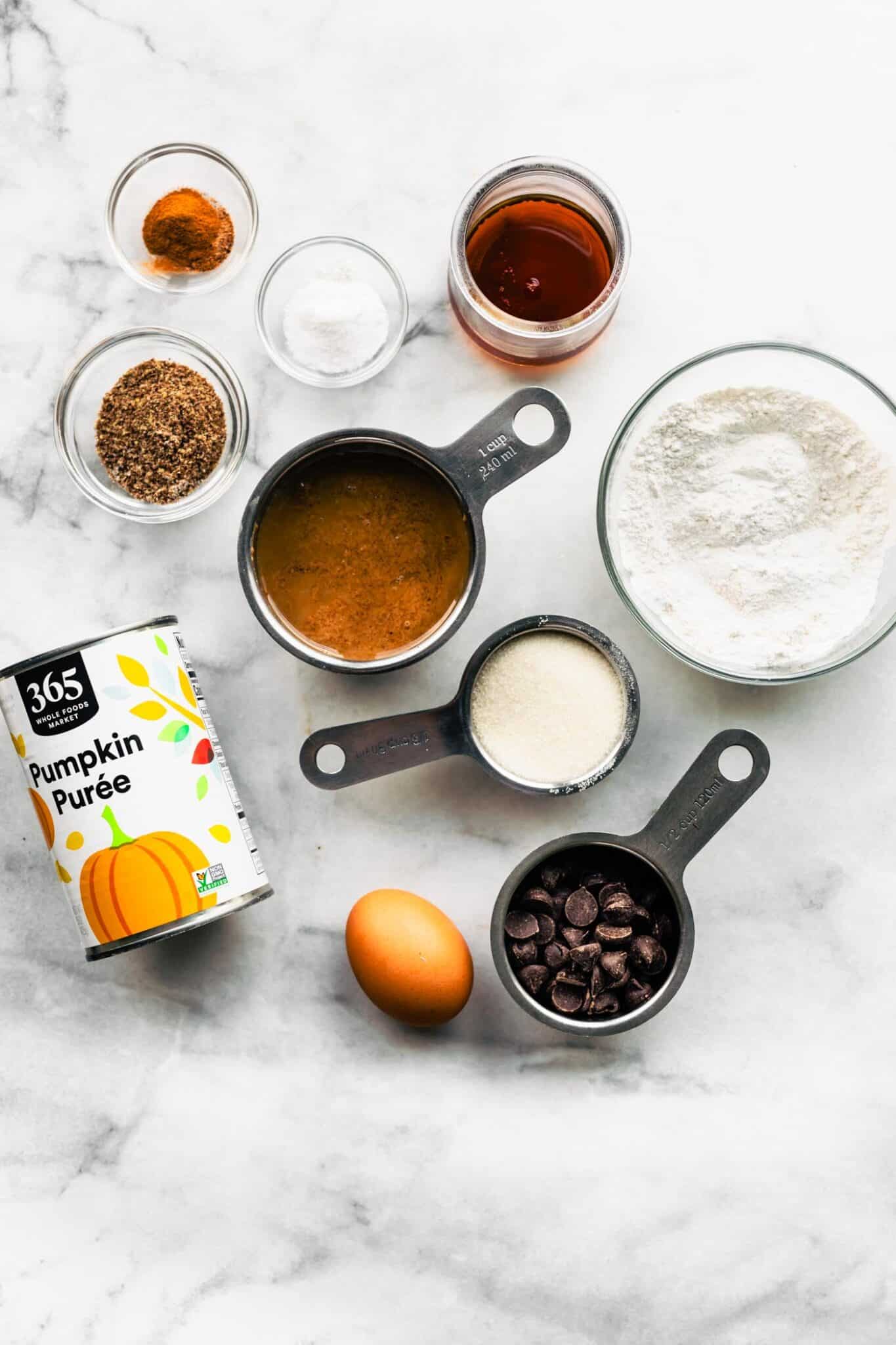 Overhead shot of pumpkin cookie ingredients in bowls and measuring cups, featuring pumpkin puree, flour, maple syrup, sugar, chocolate chips, flaxseed, egg, and spices.