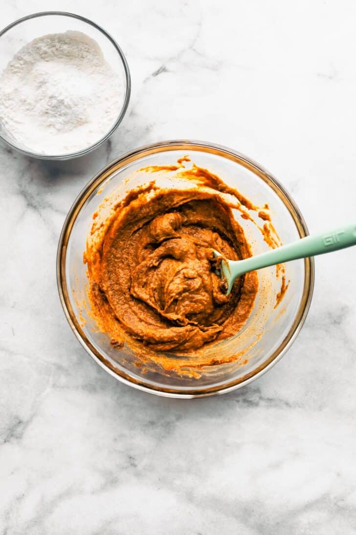 Glass bowl of pumpkin cookie wet ingredients mixed together with a spatula, with a bowl of flour nearby.