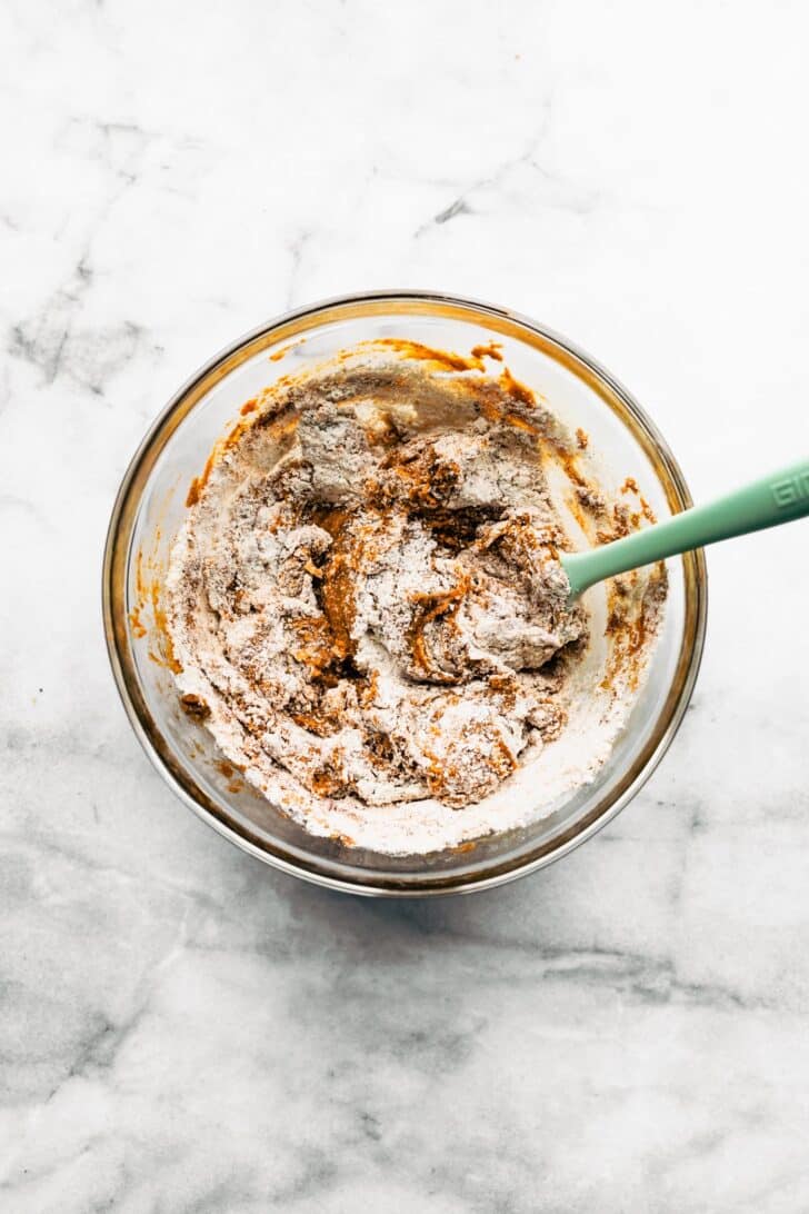 Pumpkin cookie batter being mixed with flour in a glass bowl, showing a partially combined texture.