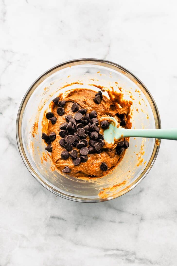 Pumpkin cookie batter in a glass bowl with chocolate chips folded in using a spatula.