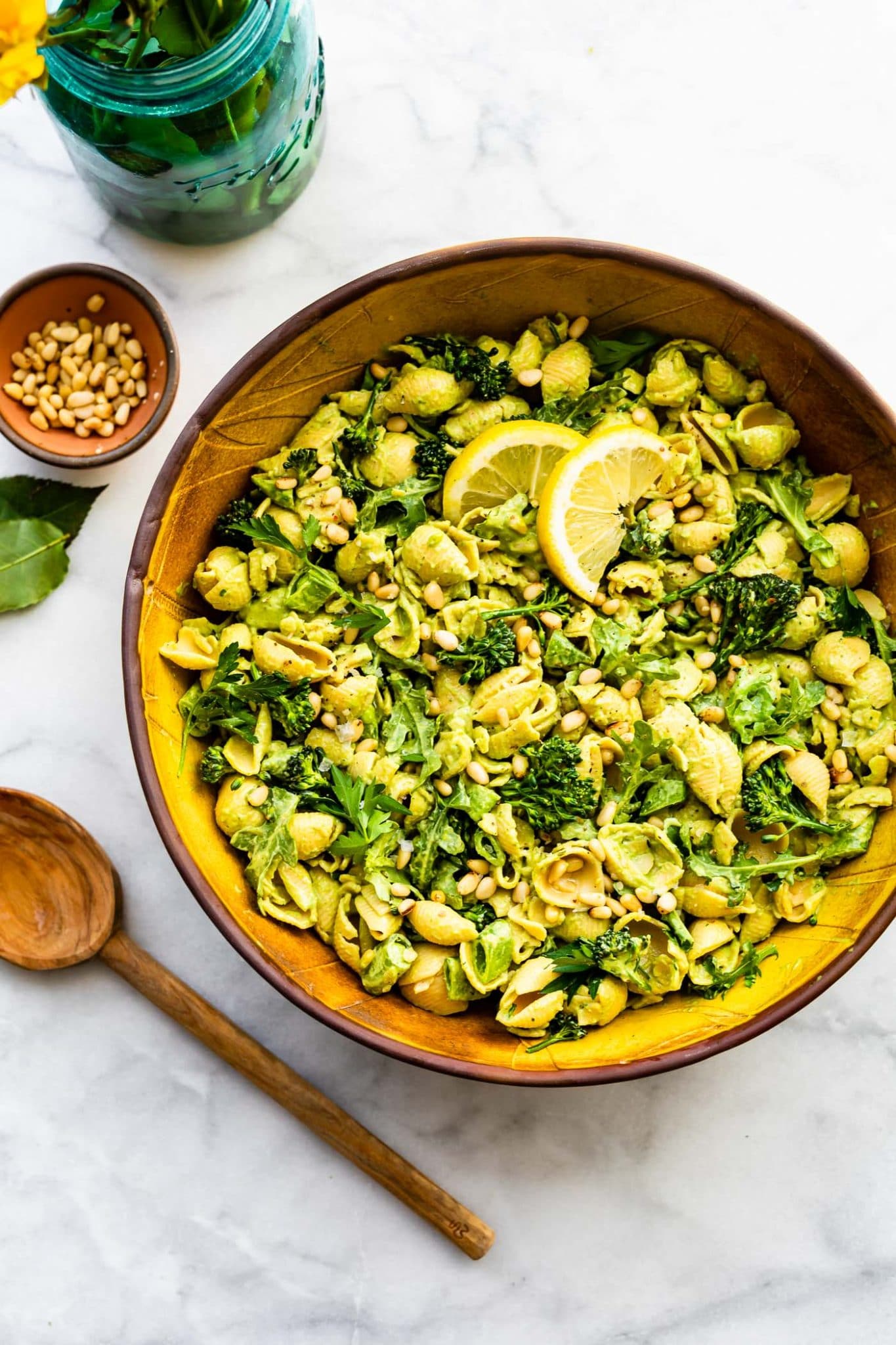 Overhead image of vegan green goddess pasta salad with a wooden spoon and flowers.