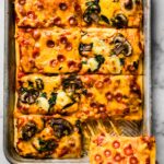 A sheet pan pizza cut into eight slices with a woman pulling a corner piece out.