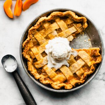Apricot pie with a slice removed with a scoop of ice cream on top; sliced apricots and ice cream scoop beside the pie.