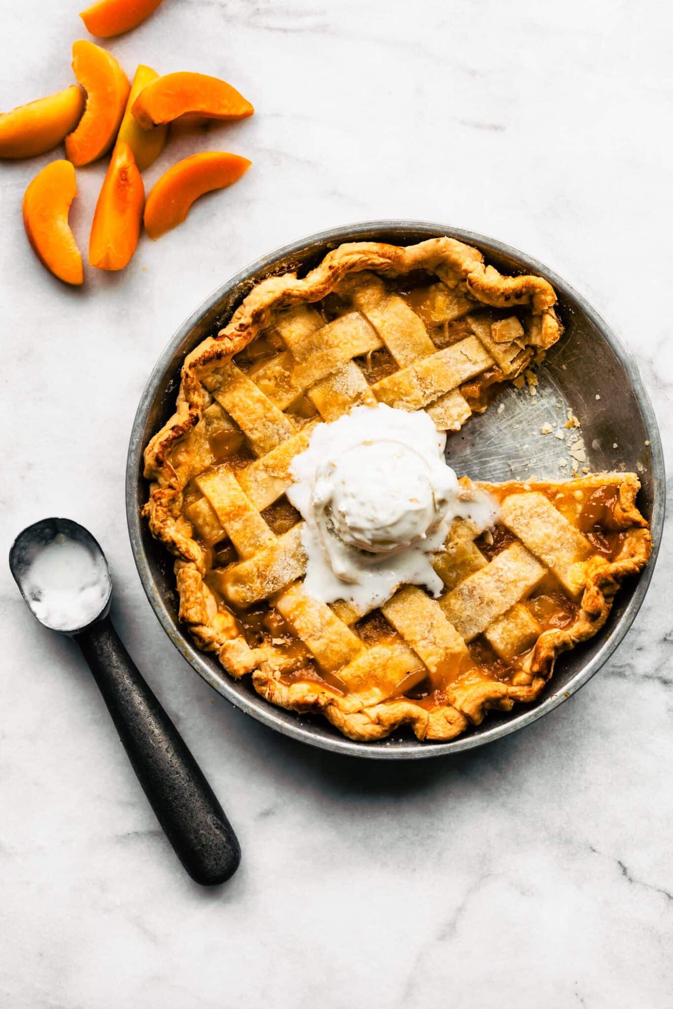 Apricot pie with a slice removed with a scoop of ice cream on top; sliced apricots and ice cream scoop beside the pie.