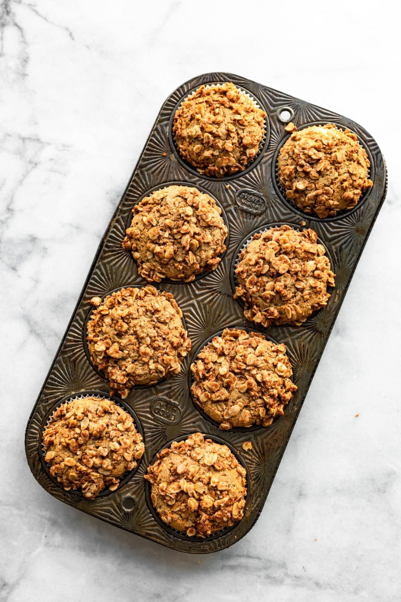 Eight baked muffins with oat crumb topping in a muffin pan.