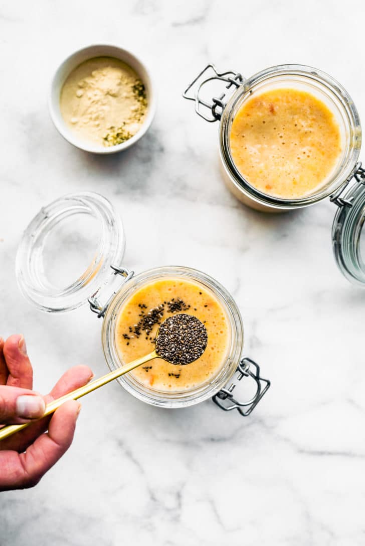 overhead image of two open glass jars of chia pudding with a woman's hand pouring chia seeds into one and a small bowl of protein in the background