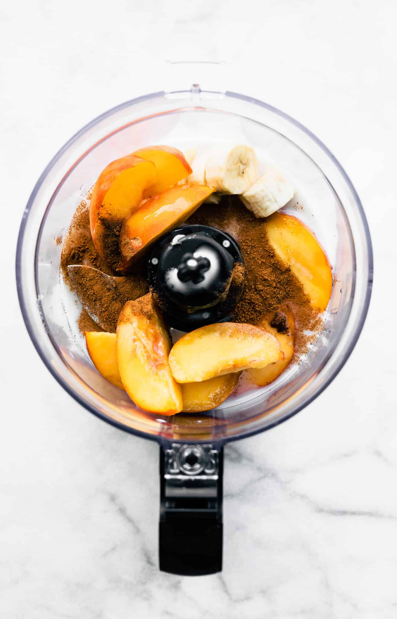 overhead image of bananas, peaches, dairy-free milk, and cinnamon in a food processor