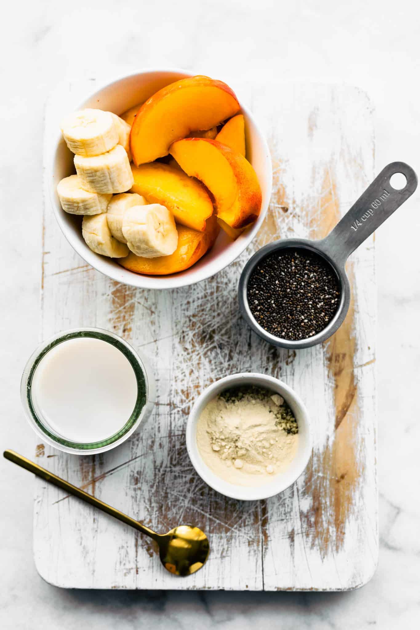 banana slices and peach slices in a white bowl, 1/4 cup full of chia seeds, protein powder and hemp seeds in a small white bowl, and dairy-free milk on a cutting board with a golden spoon to create chia pudding