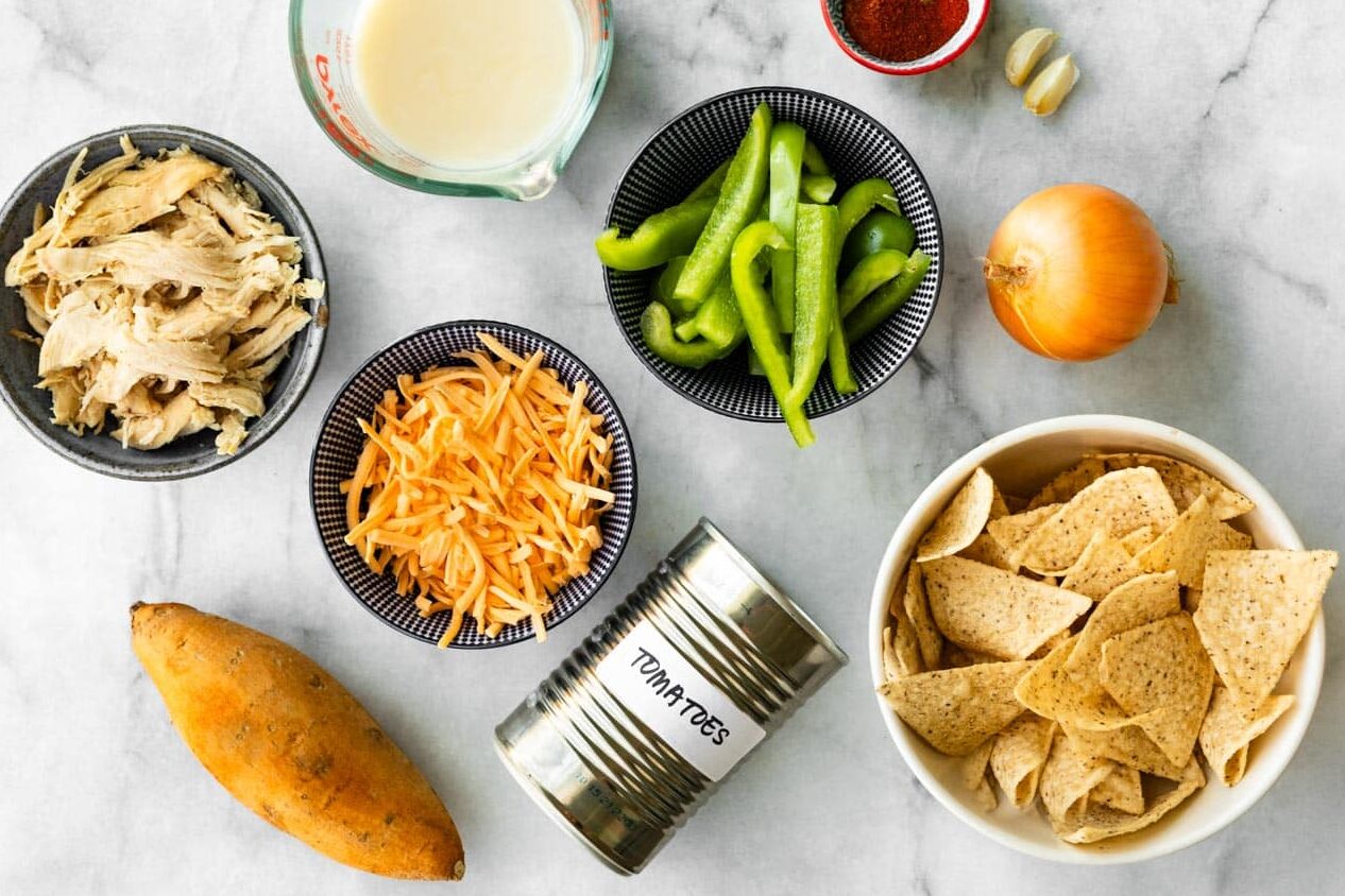 Overhead view of ingredients for chicken fajita casserole, including shredded chicken, shredded cheese, green bell peppers, onion, garlic, sweet potato, canned tomatoes, tortilla chips, milk, and spices arranged on a marble surface.