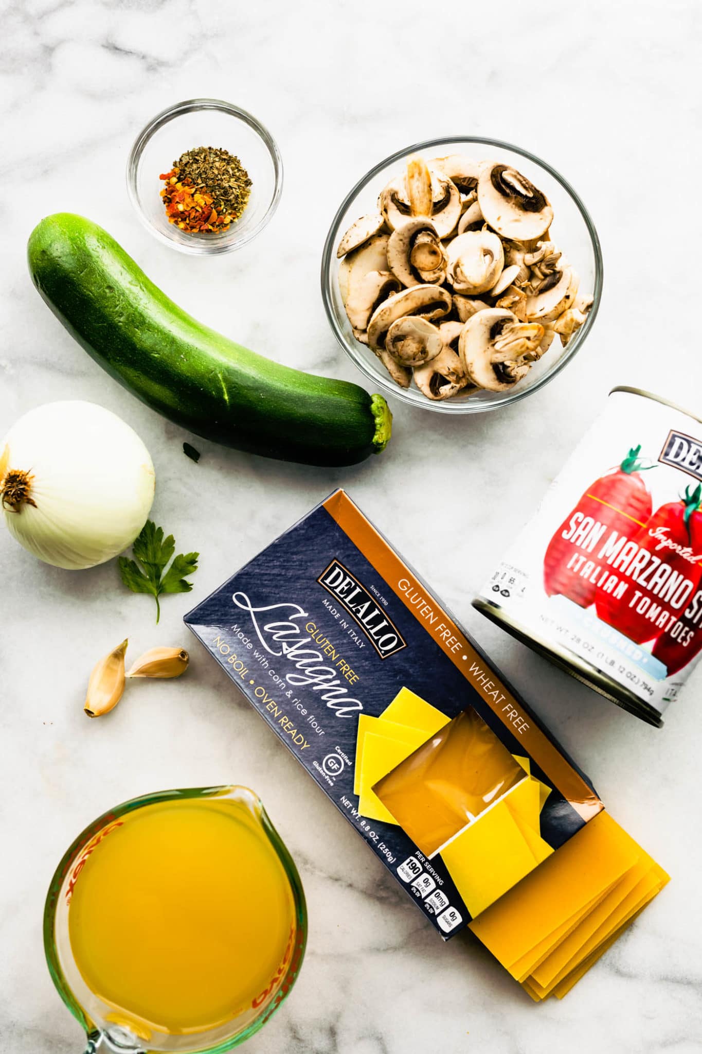 chicken broth, garlic cloves, an onion, a zucchini, spices, mushrooms, Italian tomatoes, and Delallo gluten-free lasagna noodles on a counter