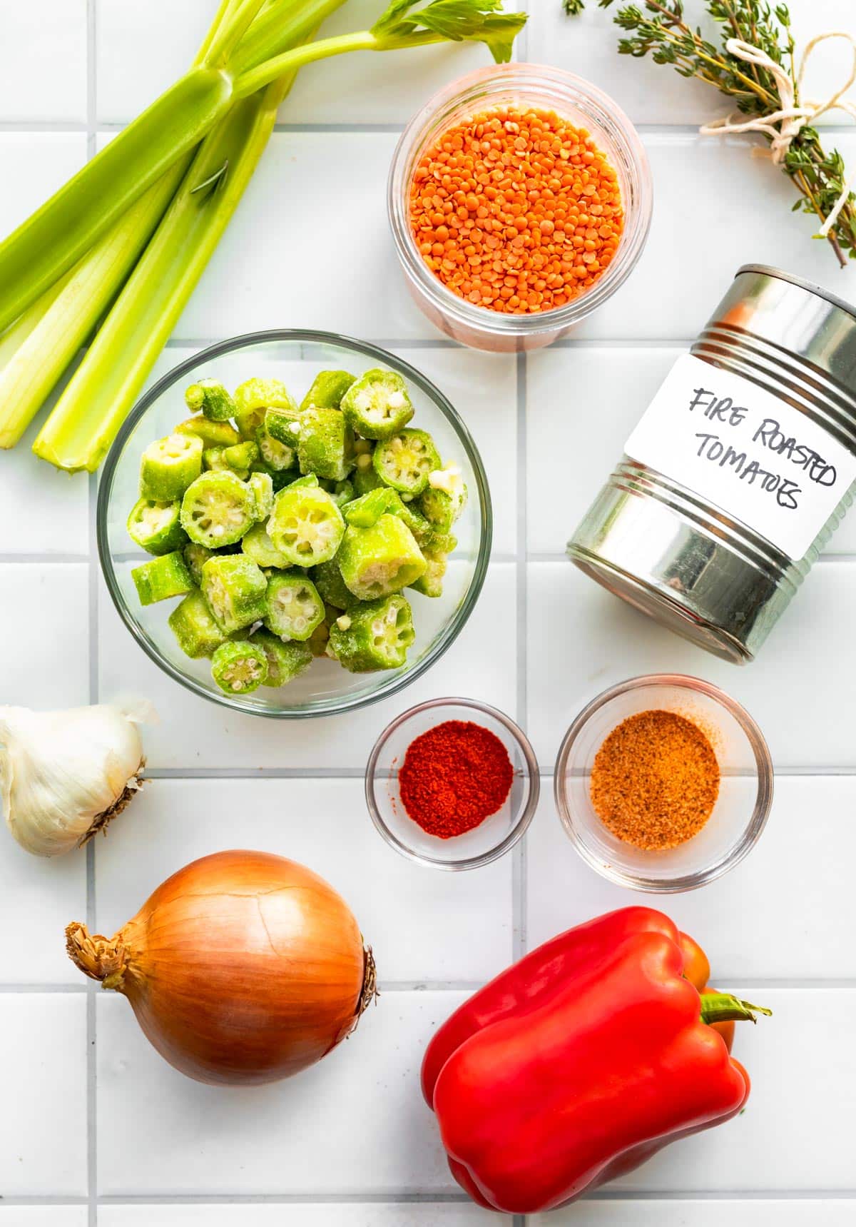 Overhead ingredient shot with okra in a glass bowl, tomatoes in a can, celery, lentils, spices, onions, garlic and red peppers.