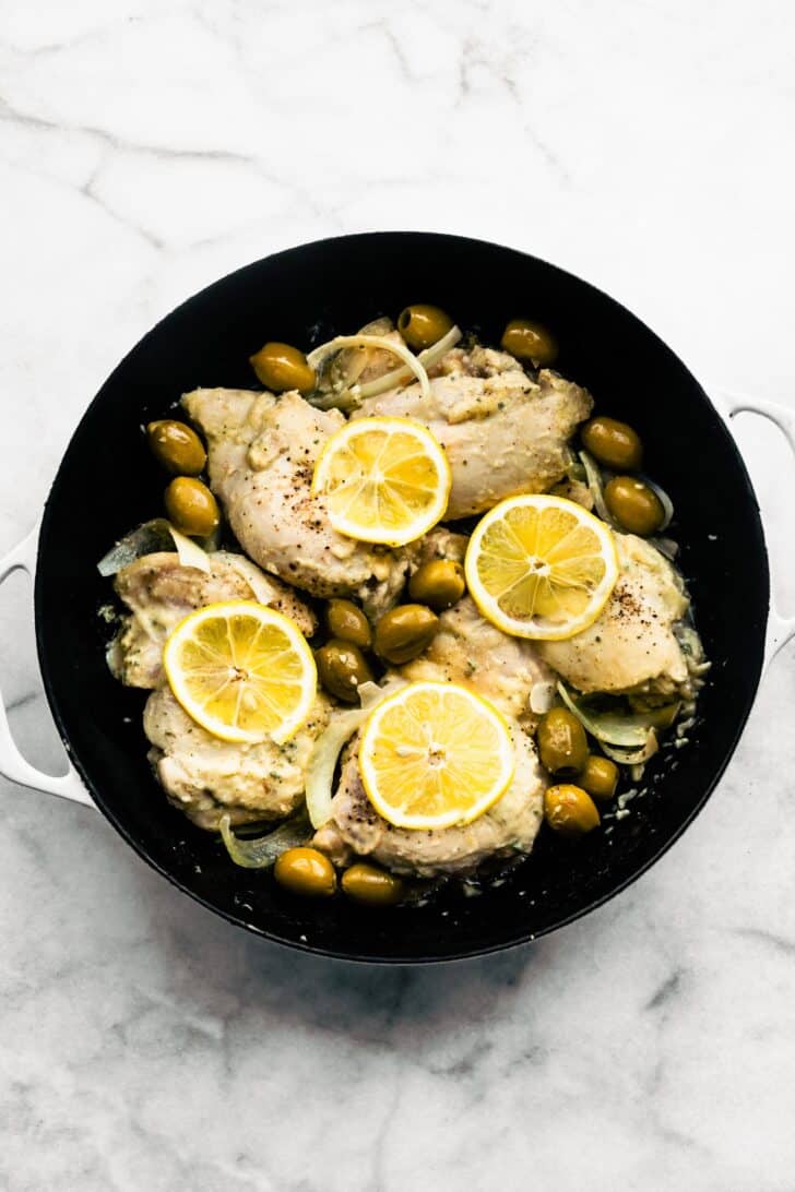Four chicken thighs browned in a skillet with lemon slices, onion and green olives.
