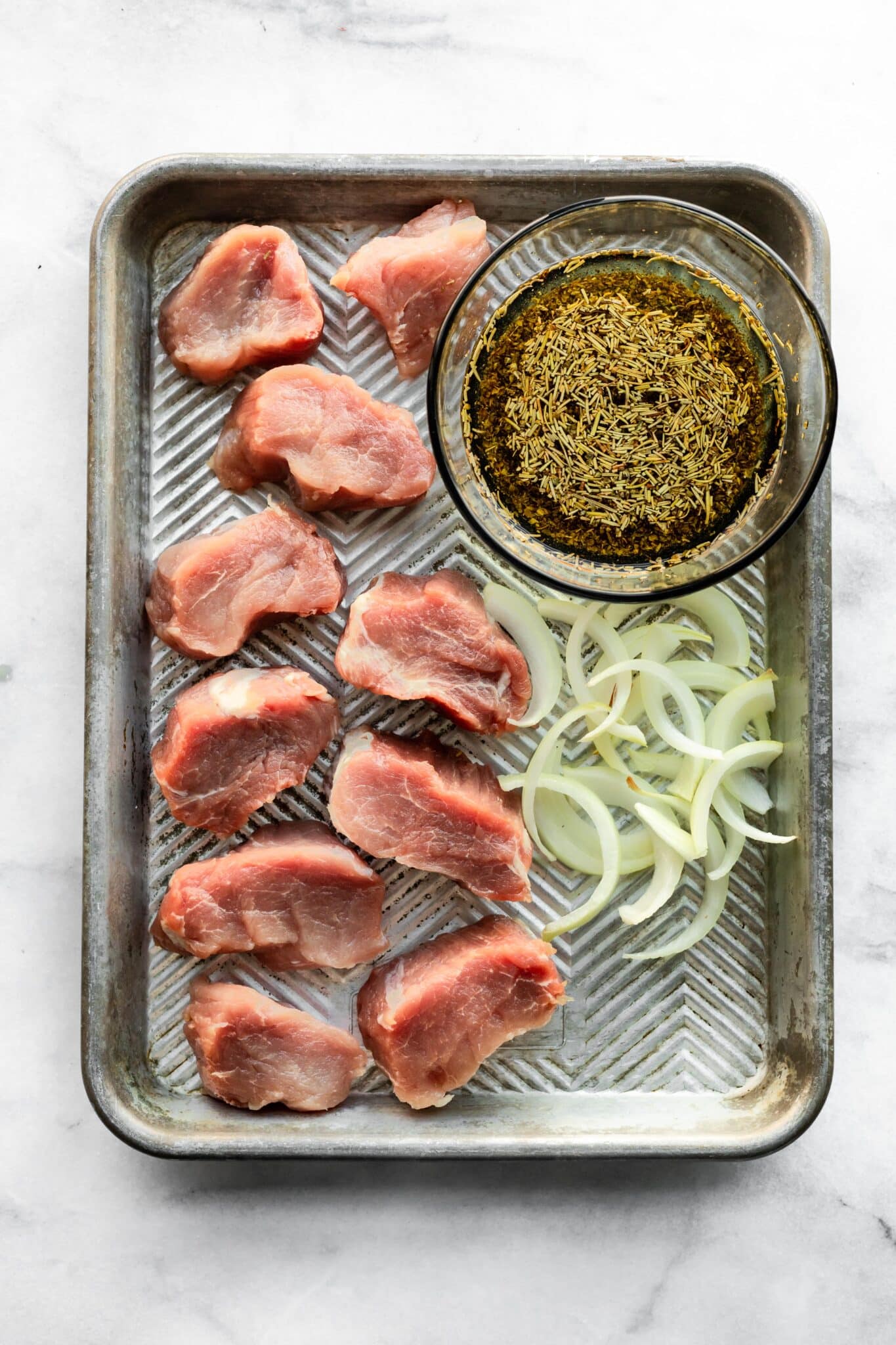 Raw pork, onions and a bowl of marinade on a metal sheet pan.